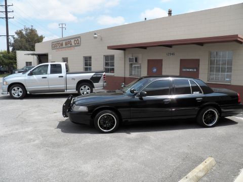 2003 Ford Crown Victoria - Side profile of lowered black Crown Victoria on black rims, silver Ram and Custom Mfg Co building behind