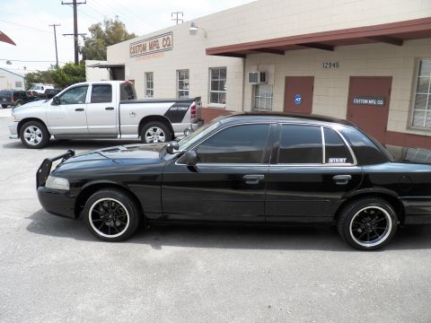 2003 Ford Crown Victoria - Lowered black Crown Victoria on black wheels sits curbside next to a silver Ram pickup outside an industrial building.