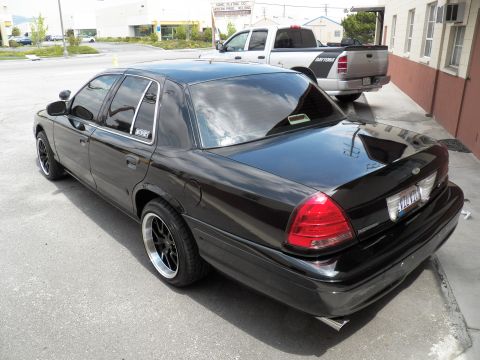 2003 Ford Crown Victoria - Black sedan shot from rear left showing tinted glass, dual exhaust and black multi-spoke wheels in a paved lot.