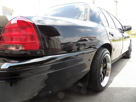 2003 Ford Crown Victoria - Glossy black sedan photographed low from the rear left, highlighting taillight and wide black rear wheel.