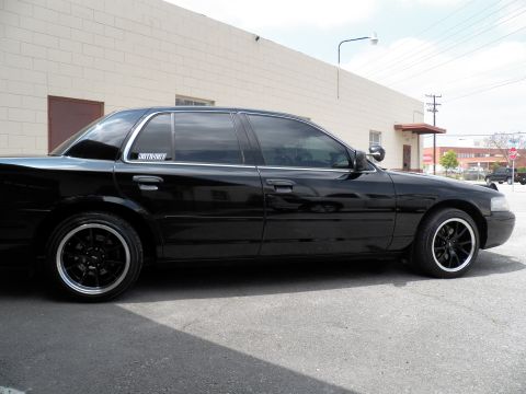 2003 Ford Crown Victoria - Full side view of a black four-door sedan, dropped over black wheels, parked beside a beige industrial wall.