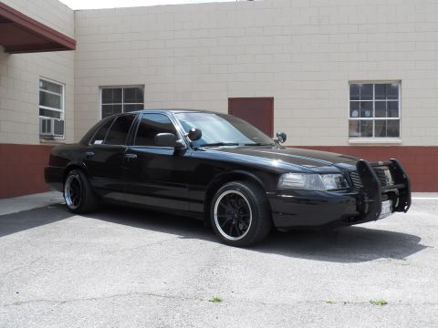 2003 Ford Crown Victoria - Black sedan front view showing push bar, spotlight and black wheels against a beige block wall.