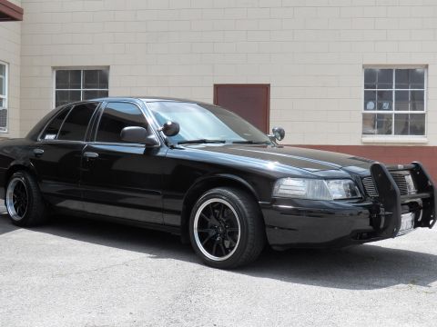2003 Ford Crown Victoria - Lowered black four-door sedan with grille guard and spotlight, photographed front-left in a paved lot.