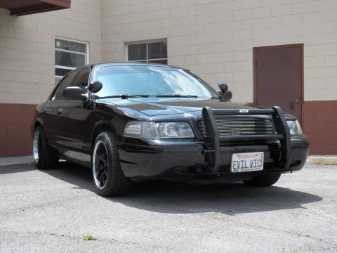 2003 Ford Crown Victoria - Black sedan shows billet grille, push bar and a California EVIL VIC plate parked at a beige building.