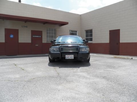 2003 Ford Crown Victoria - Distant head-on view of a black sedan with grille guard, parked before the 12946 Custom Mfg Co building.