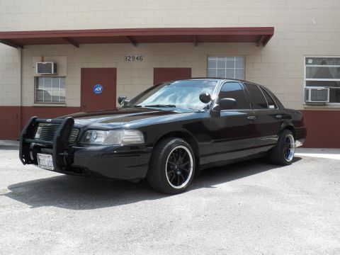 2003 Ford Crown Victoria - Lowered black sedan with push bar and pillar spotlight angled toward camera in an industrial lot.