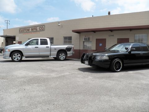 2003 Ford Crown Victoria - Silver Ram crew-cab pickup beside a black sedan with grille guard outside Custom Mfg Co Screw Machine Products.