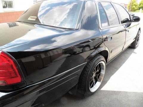 2003 Ford Crown Victoria - Glossy black Ford Crown Victoria seen from the rear quarter, showing dark aftermarket wheels.