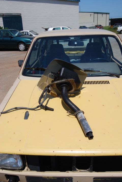 1975 Ford Ranchero - A detached fuel filler neck assembly rests on the hood of a yellow car, with other vehicles in the background.