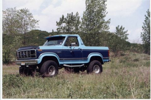 1985 Ford Bronco - This two-tone blue Ford Bronco is captured in a three-quarter front view, parked amidst tall grass and trees.