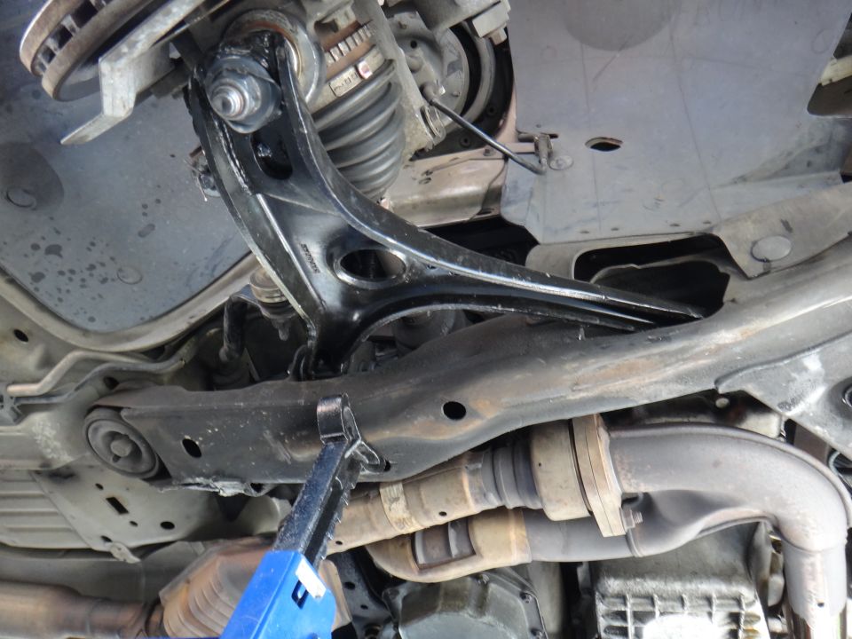 Close-up of a car's front suspension control arm and ball joint assembly, with exhaust components visible below