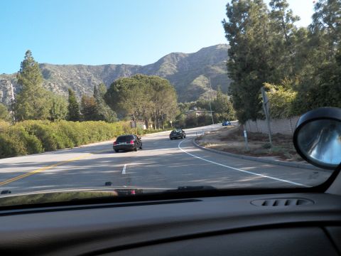 2003 Ford Crown Victoria - Driver POV through a windshield: a winding tree-lined mountain road with a dark car ahead.