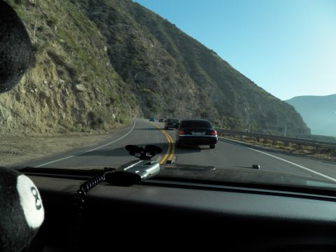 2003 Ford Crown Victoria - Driver POV down a guardrail-lined mountain road, dark sedan ahead and a fuzzy dice on dash.