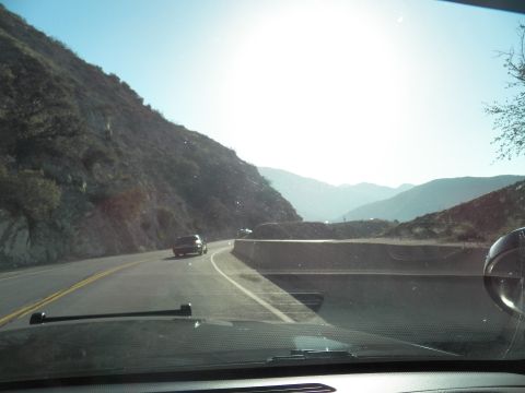 2003 Ford Crown Victoria - Backlit canyon road bends downhill past rocky slopes with a dark car ahead through the glass.