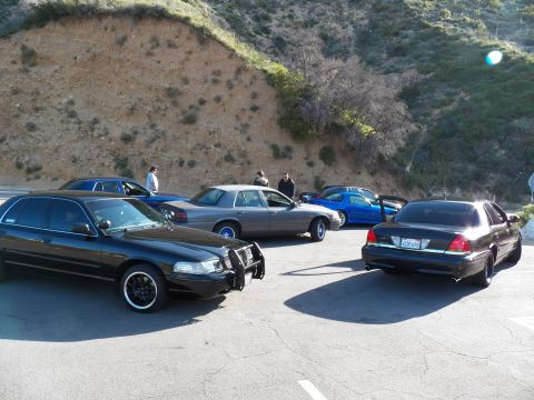 2003 Ford Crown Victoria - Cluster of dark sedans plus a blue car at a canyon pullout, people gathered between them.