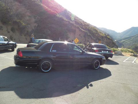 2003 Ford Crown Victoria - Rear-quarter of a black sedan at a sunlit mountain pullout, more cars and people nearby.