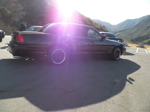 2003 Ford Crown Victoria - Rear three-quarter view of a black sedan on chrome-lipped wheels in a mountain parking area at sunset