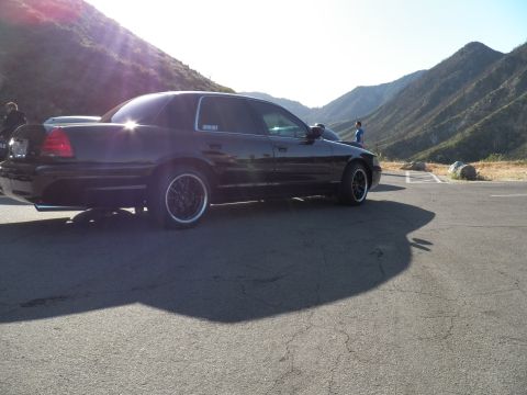 2003 Ford Crown Victoria - Side-rear view of a black sedan with dark wheels, mountain canyon and bystanders behind