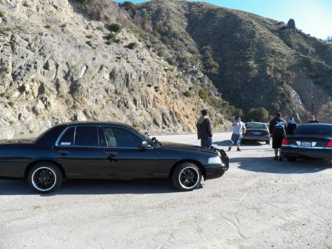 2003 Ford Crown Victoria - Profile of a black sedan on black wheels in a dirt mountain pullout among a group of cars