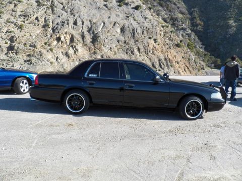 2003 Ford Crown Victoria - Side view of a lowered black sedan parked in a dirt pullout, blue sedan and people nearby