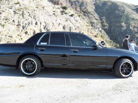 2003 Ford Crown Victoria - Passenger-side view of a black sedan parked on gravel below a rocky ridge, people at right