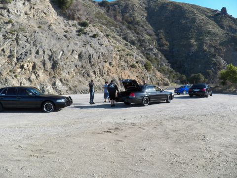 2003 Ford Crown Victoria - Several dark full-size sedans gathered in a gravel pullout below a steep rock face