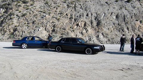 2003 Ford Crown Victoria - Two full-size sedans and a small group of people in a gravel pullout beneath a rocky slope
