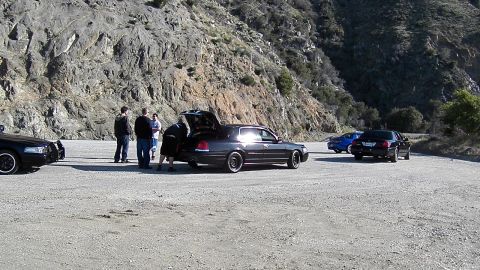 2003 Ford Crown Victoria - Group gathered behind a black sedan with open trunk in a dirt pullout, other cars around