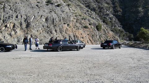 2003 Ford Crown Victoria - Wide view of several dark sedans and bystanders in a dirt pullout below a rocky ridge