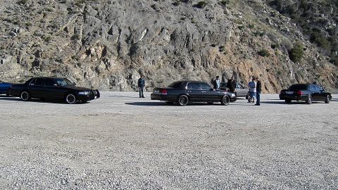 2003 Ford Crown Victoria - Several dark sedans and people lined up in a dirt mountain pullout below a rocky face