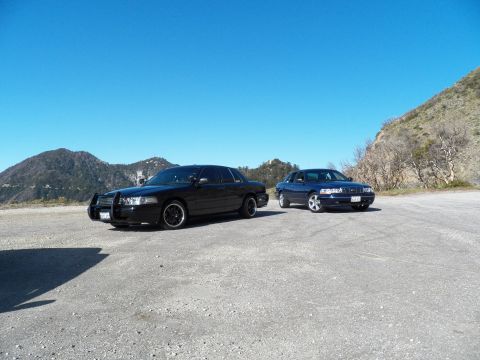 2003 Ford Crown Victoria - Front-quarter view of a black sedan beside a blue sedan in a gravel pullout, mountains behind