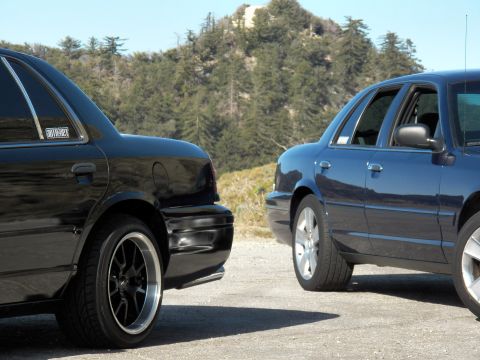 2003 Ford Crown Victoria - Close rear-quarter view of a black and a blue full-size sedan with mountain pines behind