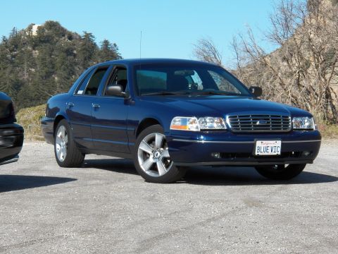 2003 Ford Crown Victoria - Ford-badged blue sedan on polished wheels parked beside bare hillside trees