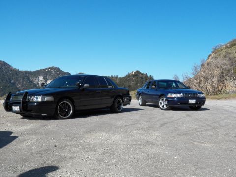 2003 Ford Crown Victoria - Two full-size sedans angled apart in a mountain clearing, one fitted with grille guard