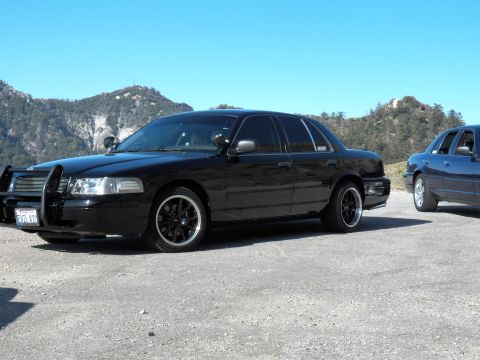 2003 Ford Crown Victoria - Black sedan on dark wheels with grille guard parked on mountain lot, blue car at right