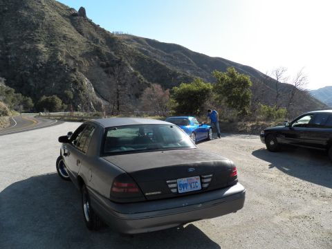 2003 Ford Crown Victoria - Rear of gray Ford sedan on a mountain road shoulder, blue and black cars ahead