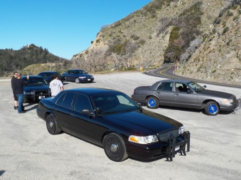 2003 Ford Crown Victoria - Black sedan with grille guard near gray sedan on bright blue wheels, people at left