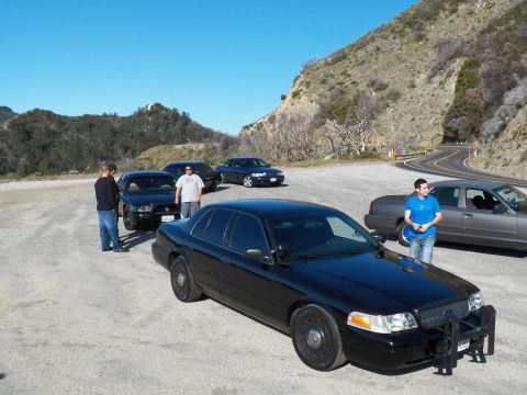 2003 Ford Crown Victoria - Black full-size sedan front three-quarter with onlookers on a sunny gravel pullout