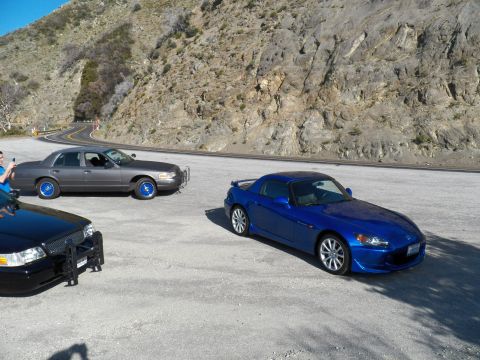 2003 Ford Crown Victoria - Blue convertible roadster parked near gray full-size sedan on a curving mountain road