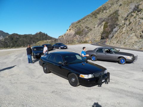 2003 Ford Crown Victoria - Black full-size sedan with grille guard, gray sedan at right and onlookers at left
