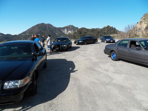 2003 Ford Crown Victoria - Black, gray and blue sedans lined up on a wide dirt pullout with peaks behind