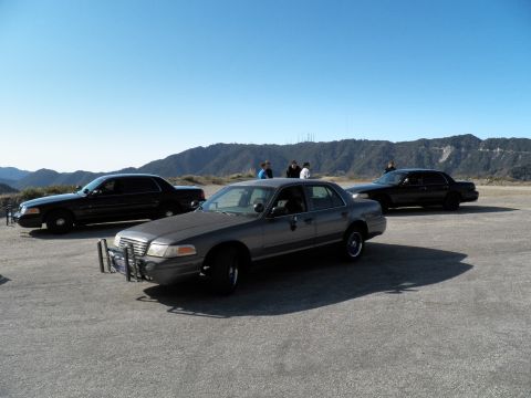 2003 Ford Crown Victoria - Gray full-size sedan parked between black sedans on a dirt lot below distant ridge