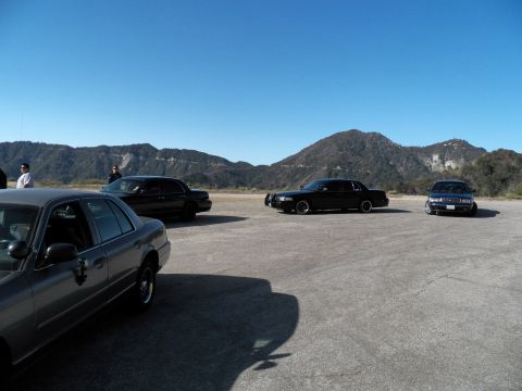 2003 Ford Crown Victoria - Several full-size sedans parked apart on a wide gravel pullout with people at left