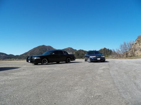 2003 Ford Crown Victoria - Pair of dark sedans on a broad gravel pullout backed by rolling mountain hills