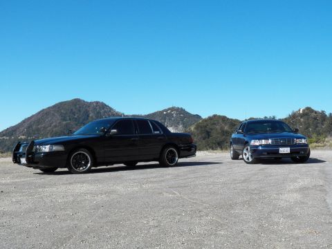 2003 Ford Crown Victoria - Black full-size sedan with grille guard beside blue Ford sedan against twin peaks