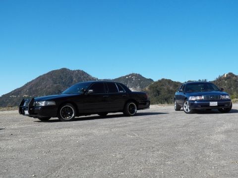 2003 Ford Crown Victoria - Black sedan with grille guard at left and blue sedan at right under clear sky