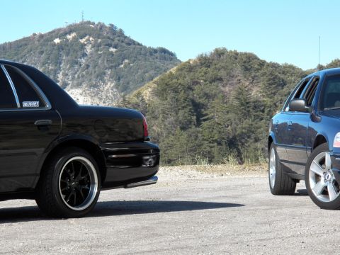 2003 Ford Crown Victoria - Tail of black full-size sedan meets blue sedan flank with forested ridge behind
