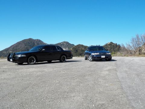 2003 Ford Crown Victoria - Pair of murdered-out full-size sedans on a dirt lot with hazy mountain ridges behind.