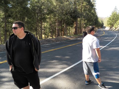 2003 Ford Crown Victoria - Two men cross a winding mountain road bordered by tall pine trees.