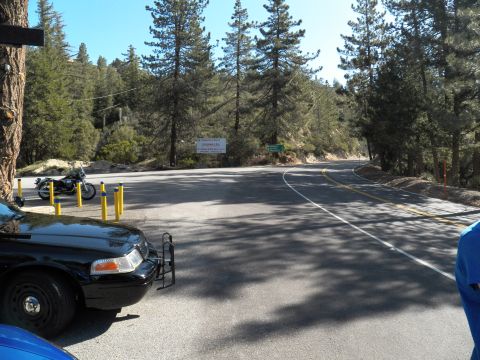 2003 Ford Crown Victoria - View across a sunlit mountain road fork, a parked motorcycle and black sedan framing the curve.